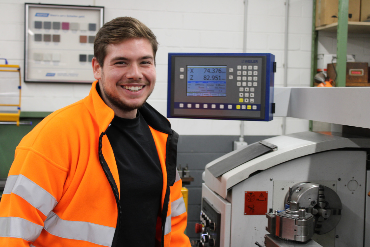 Ein dualer Studierender steht vor einer Maschine und schaut lächenld in die Kamera.