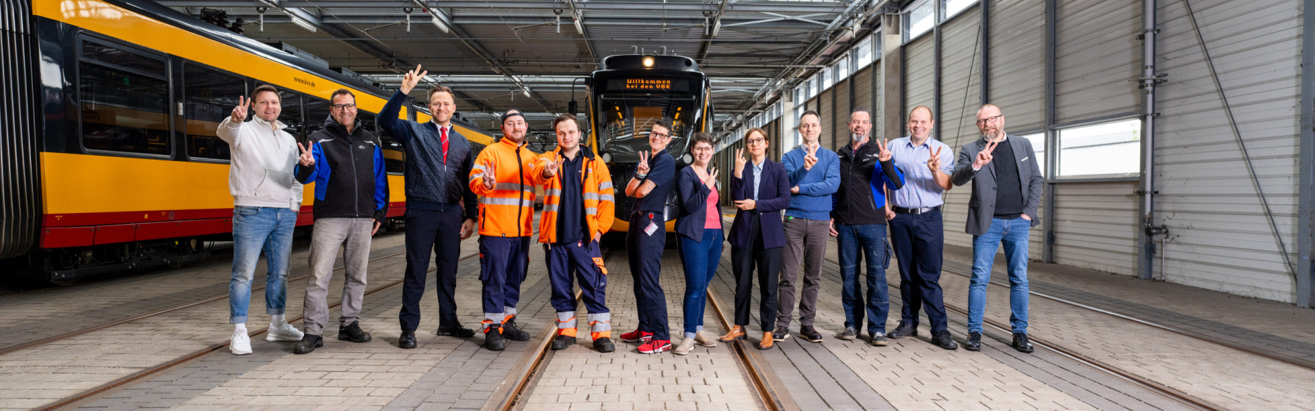 Es stehen zwölf Mitarbeitende, aus unterschiedlichen Bereichen der Unternehmen, nebeneinander mit der #TeamV Pose 