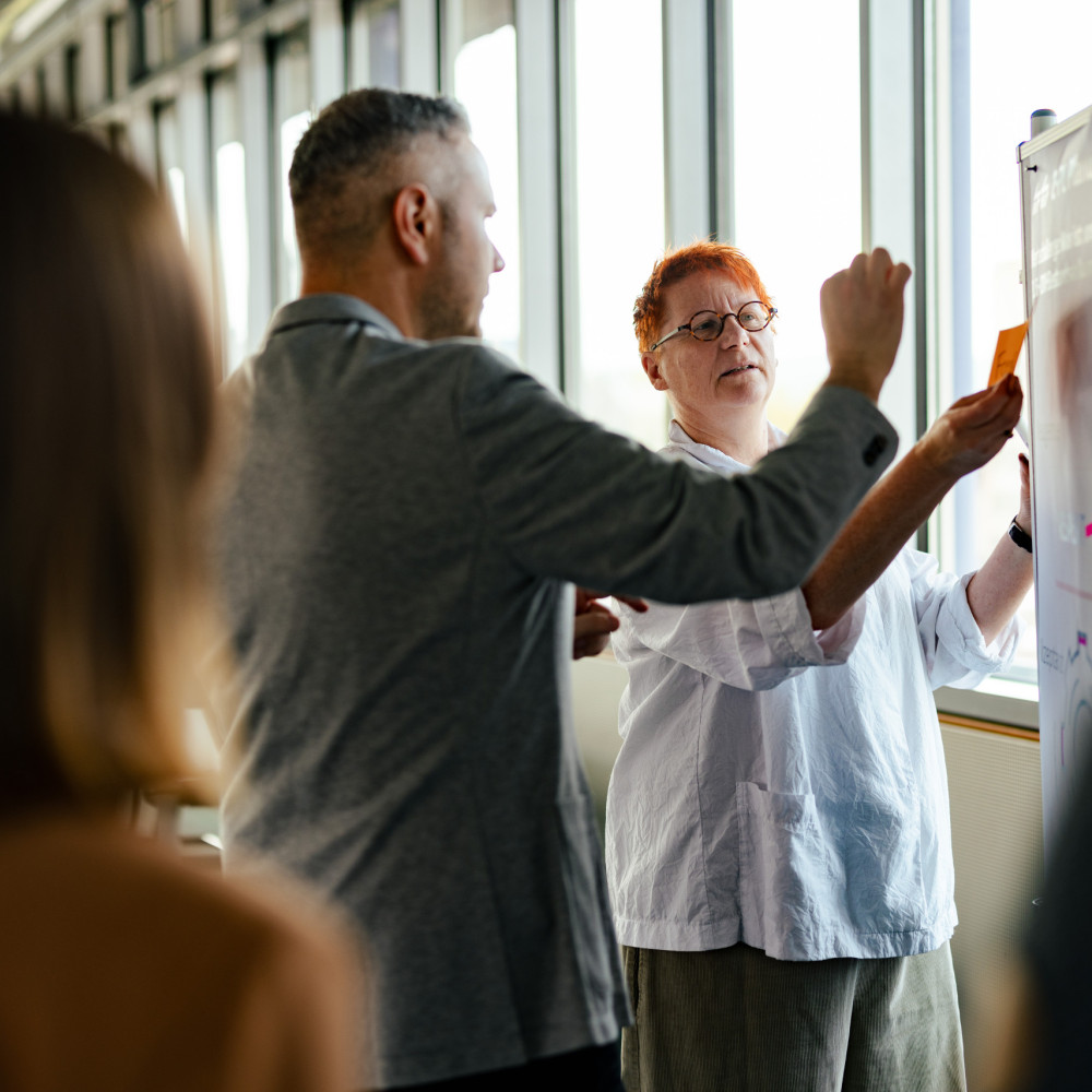 Eine Mitarbeiterin und ein Mitarbeiter besprechen etwas an einer Pinwand.