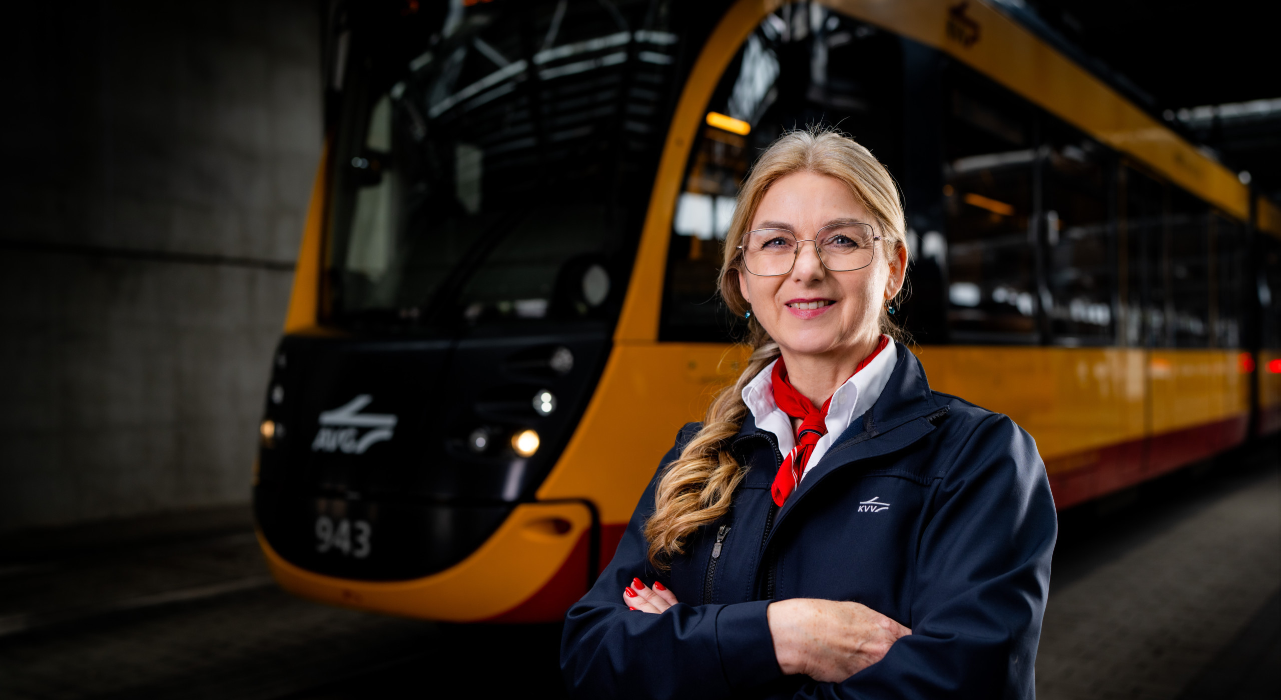 Eine freundliche Straßenbahnfahrerin mit blonden Haaren und Brille steht mit verschränkten Armen vor einer gelb-schwarzen Straßenbahn in einem überdachten Bahndepot. Sie trägt eine dunkelblaue KVV-Uniform mit rotem Halstuch und lächelt selbstbewusst in die Kamera.