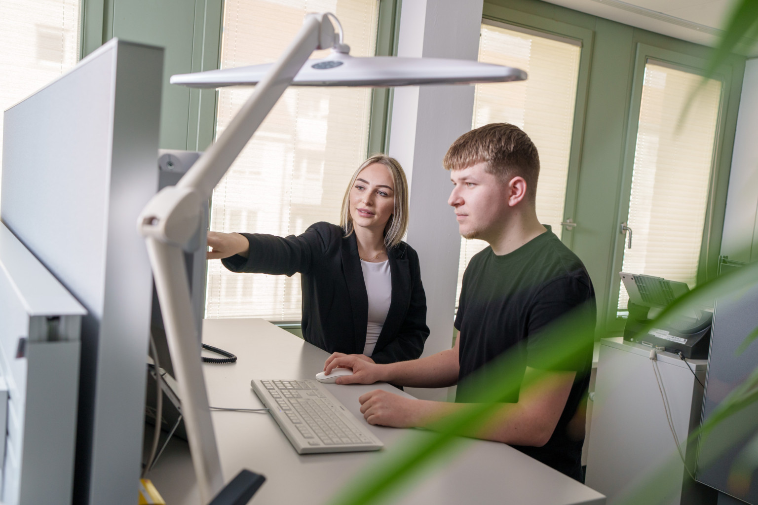 Eine Mitarbeiterin der Verwaltung steht an ihrem Arbeitsplatz und zeigt einem Azubi etwas auf dem PC-Monitor..